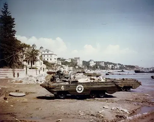 An amphibious military vehicle on a beach, Italian buildings in the background, blue sky.