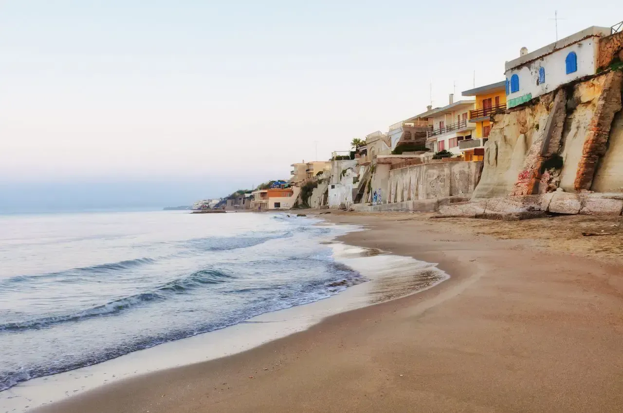 Sandy beach with gentle waves, light blue sky, and buildings on a cliff.