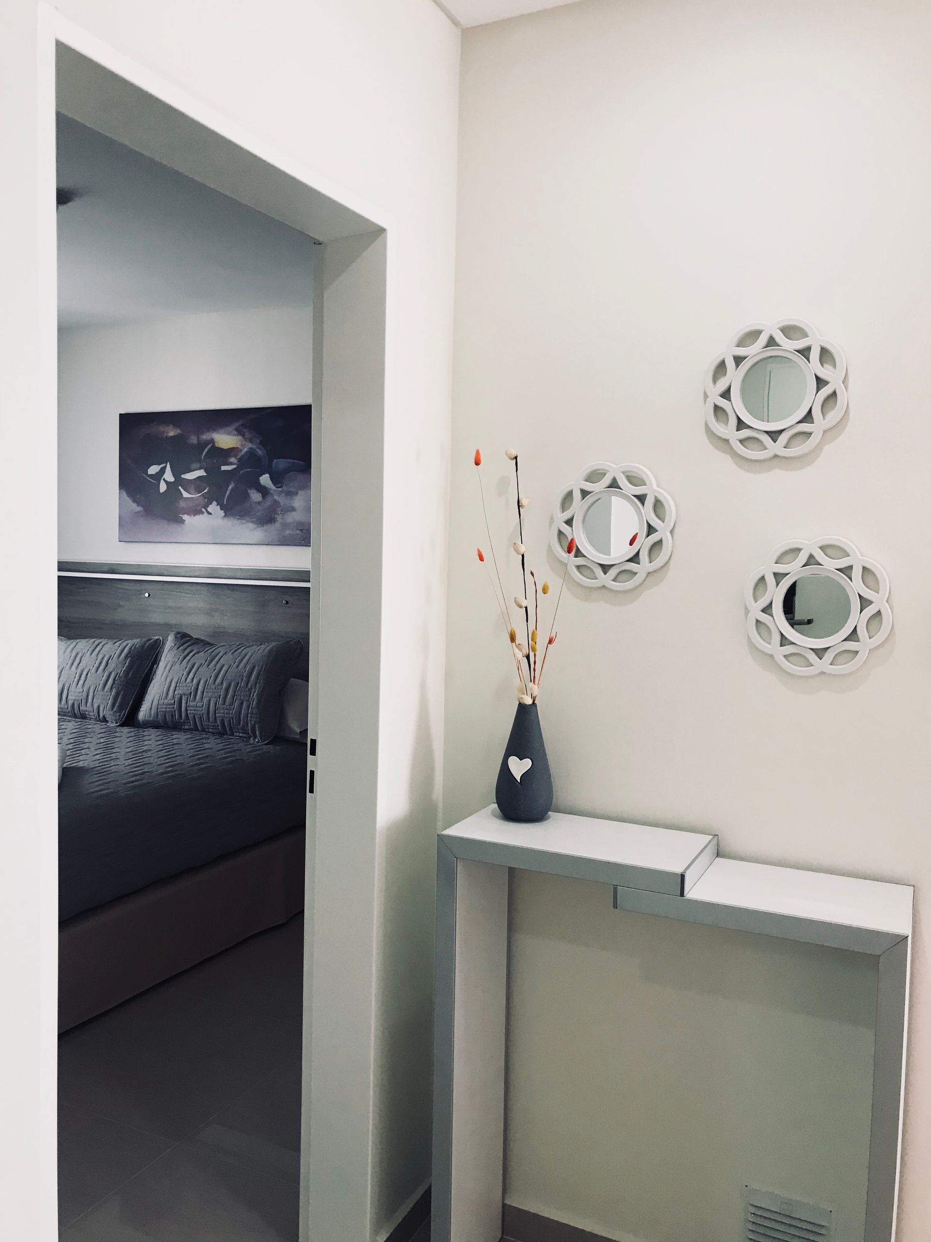 Hallway with white walls, three decorative mirrors, a shelf with a vase, and a glimpse into a bedroom.