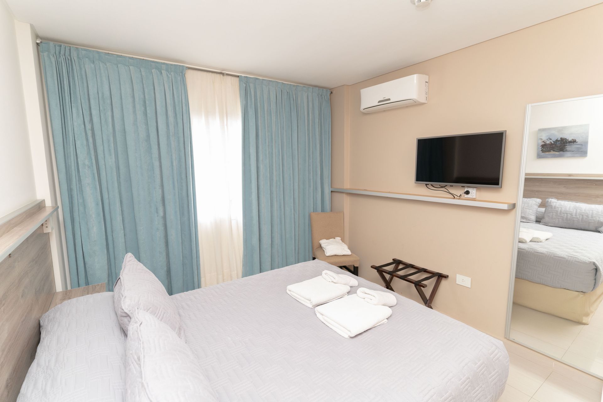 Hotel room with bed, curtains, TV, mirror. Light blue curtains, beige walls, white towels on bed.
