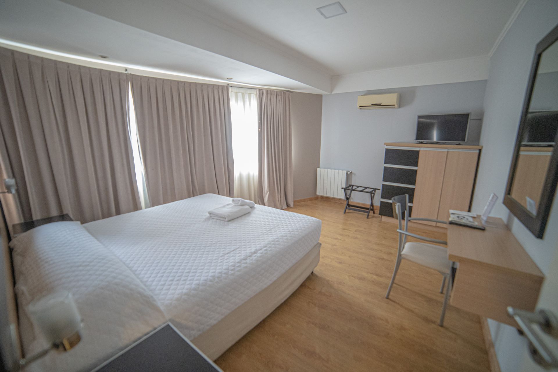 Hotel room with a bed, desk, TV, and closed curtains. Light wood floor, white bed linens, and a small AC unit.