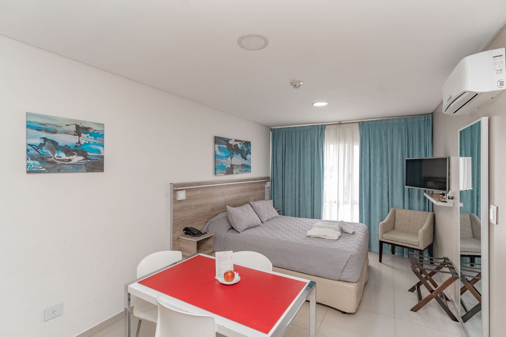 Hotel room with bed, table, chair, TV, curtains, and artwork.