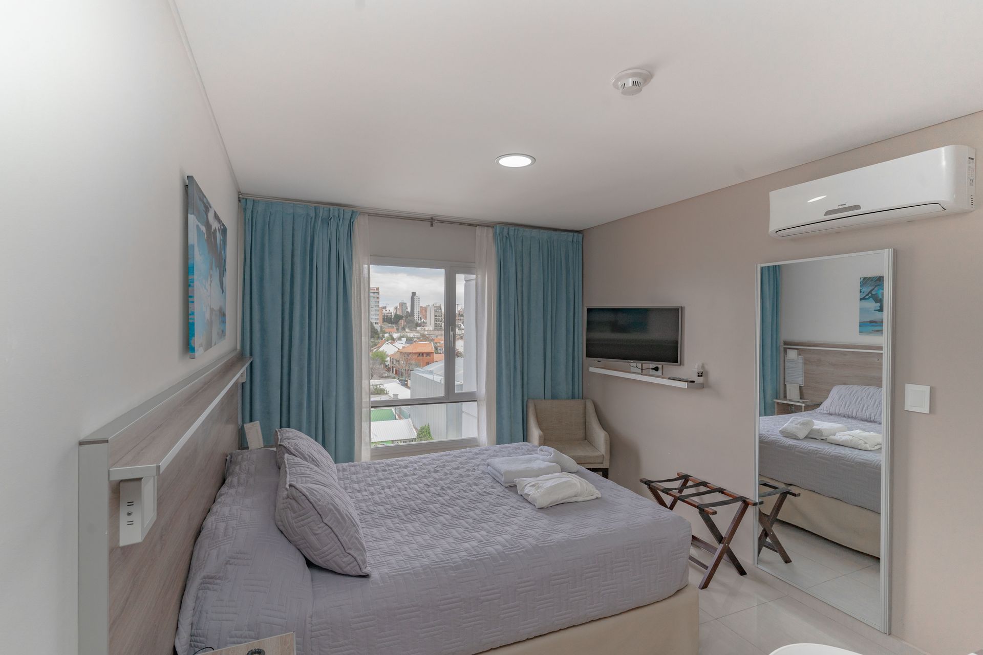 Hotel bedroom with a bed, TV, chair, and large mirror. Window with curtains and air conditioner.