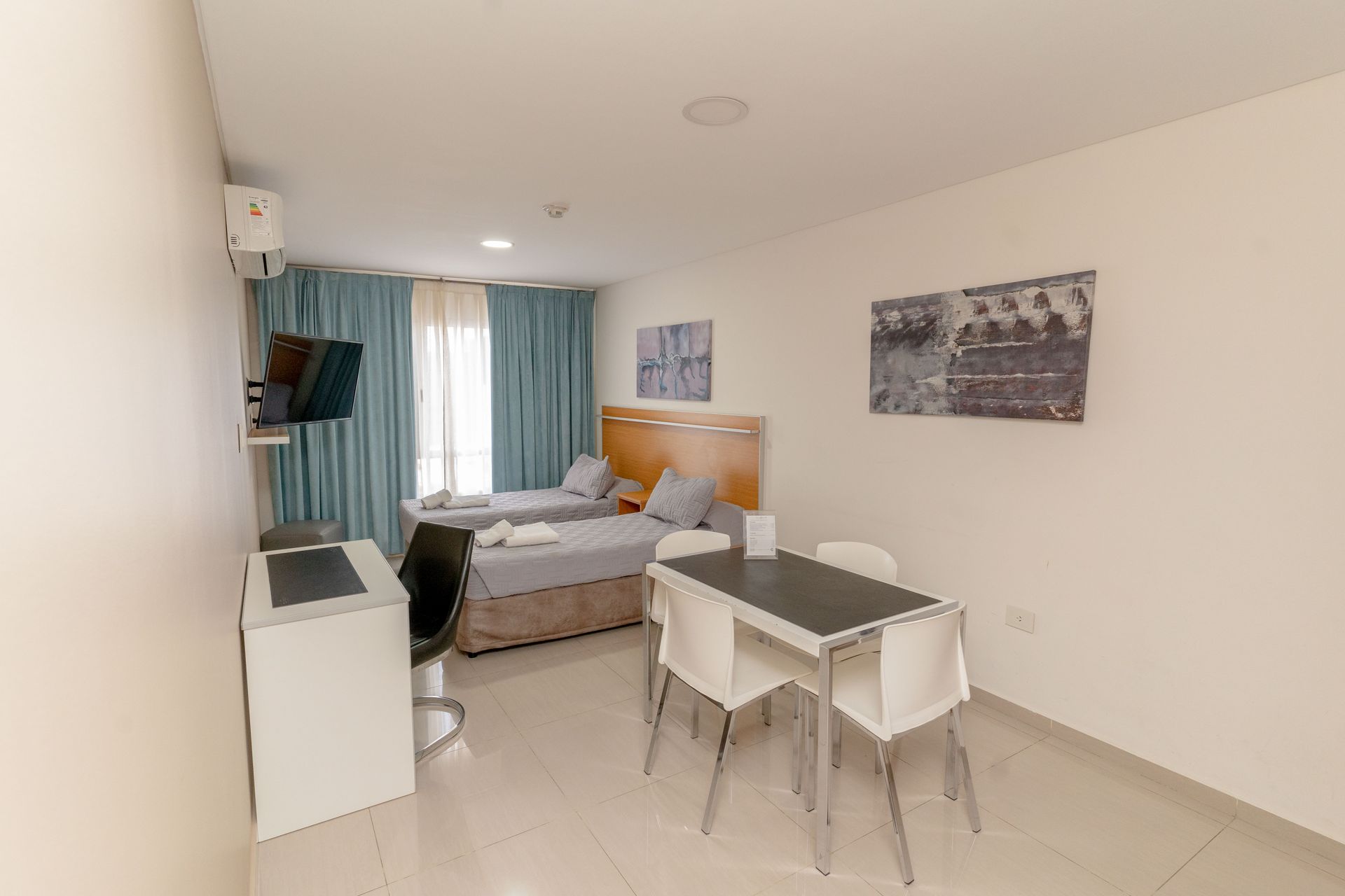 Bedroom with two beds, small dining table with chairs, TV, and window with curtains.