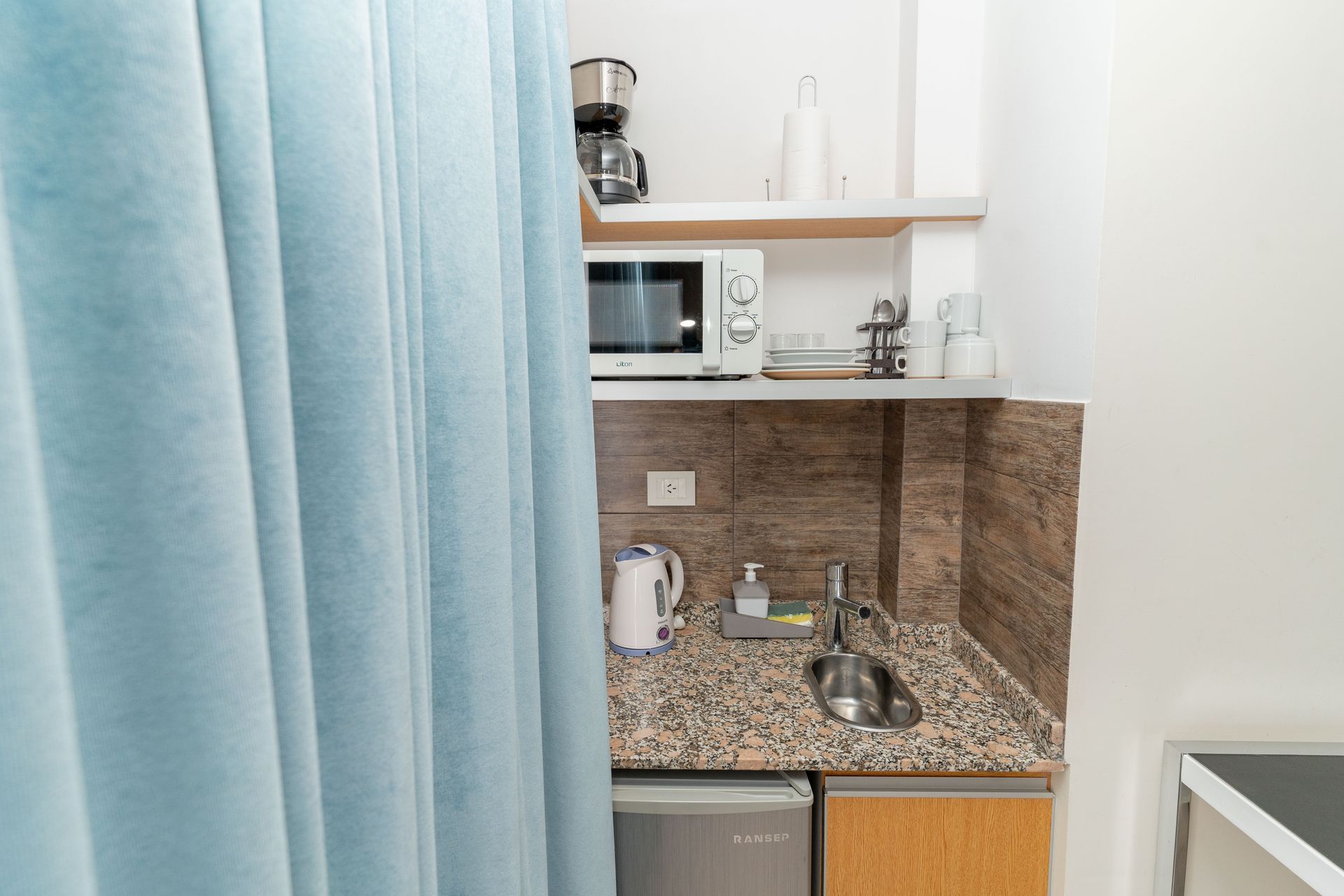 Small kitchenette with microwave, sink, and mini-fridge, partially hidden by a blue curtain.