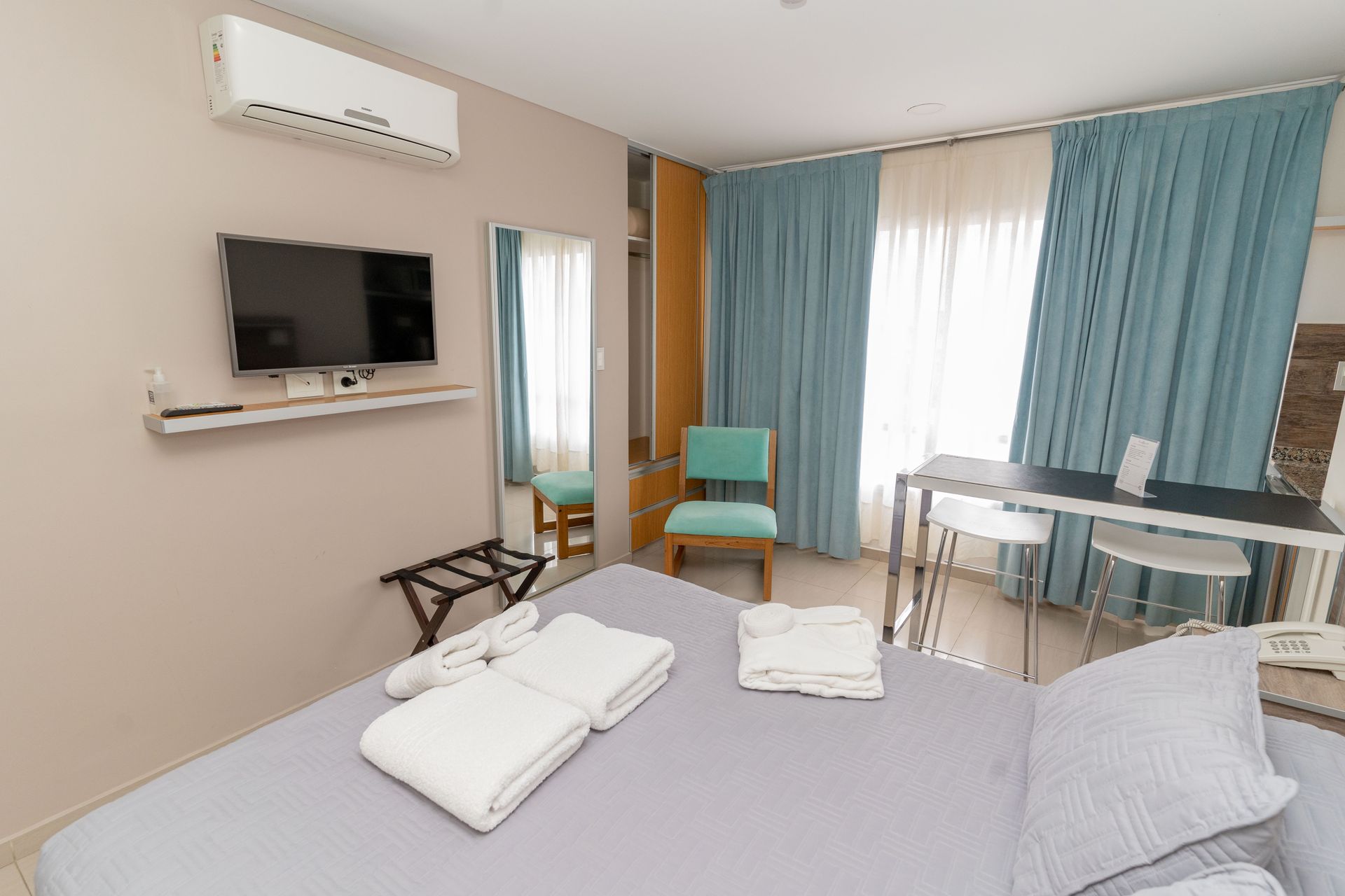 Hotel room with a bed, TV, desk, and air conditioning unit; light blue and beige color scheme.