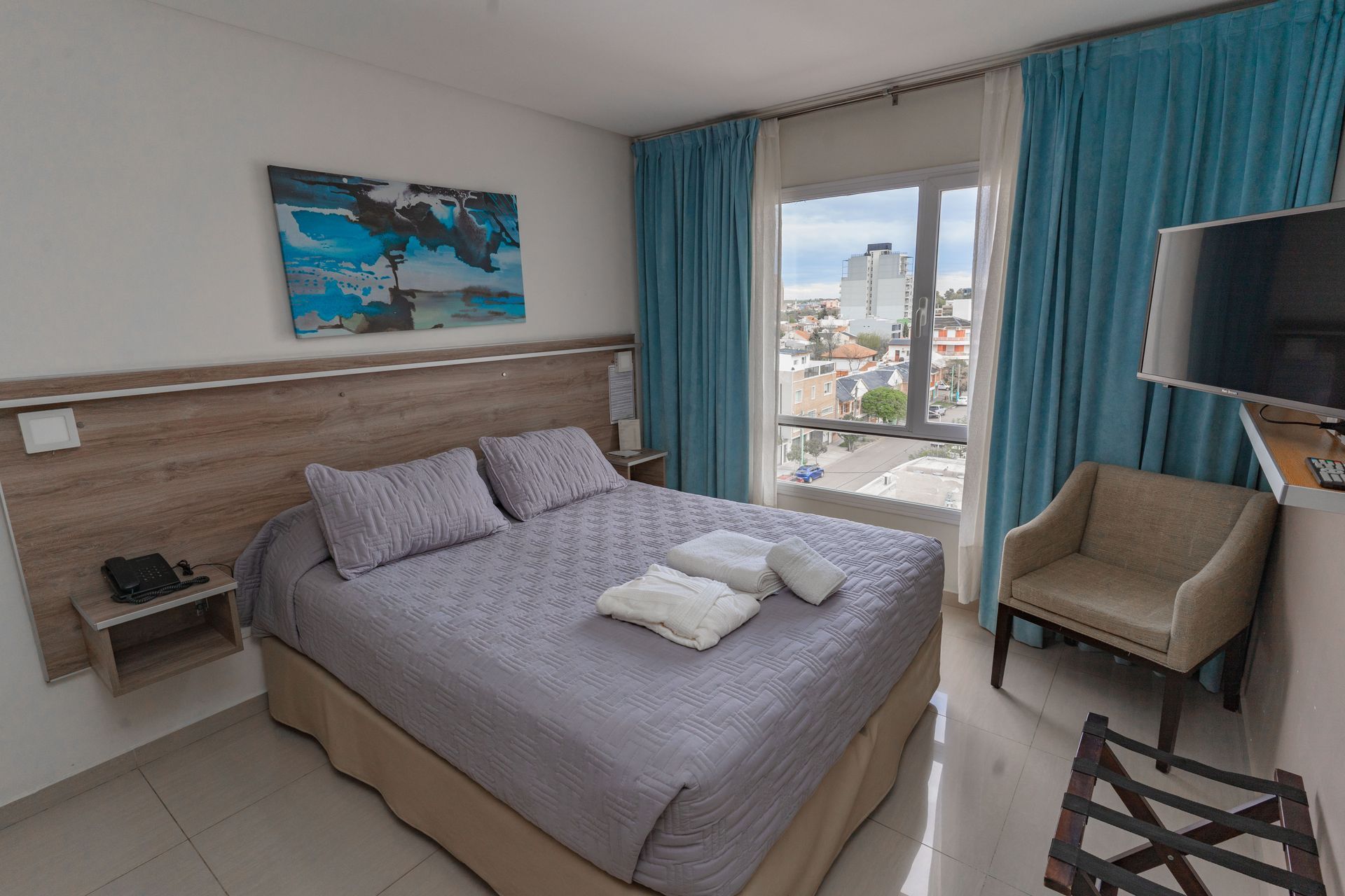 Hotel room with a bed, artwork, a chair, and a view of the city.