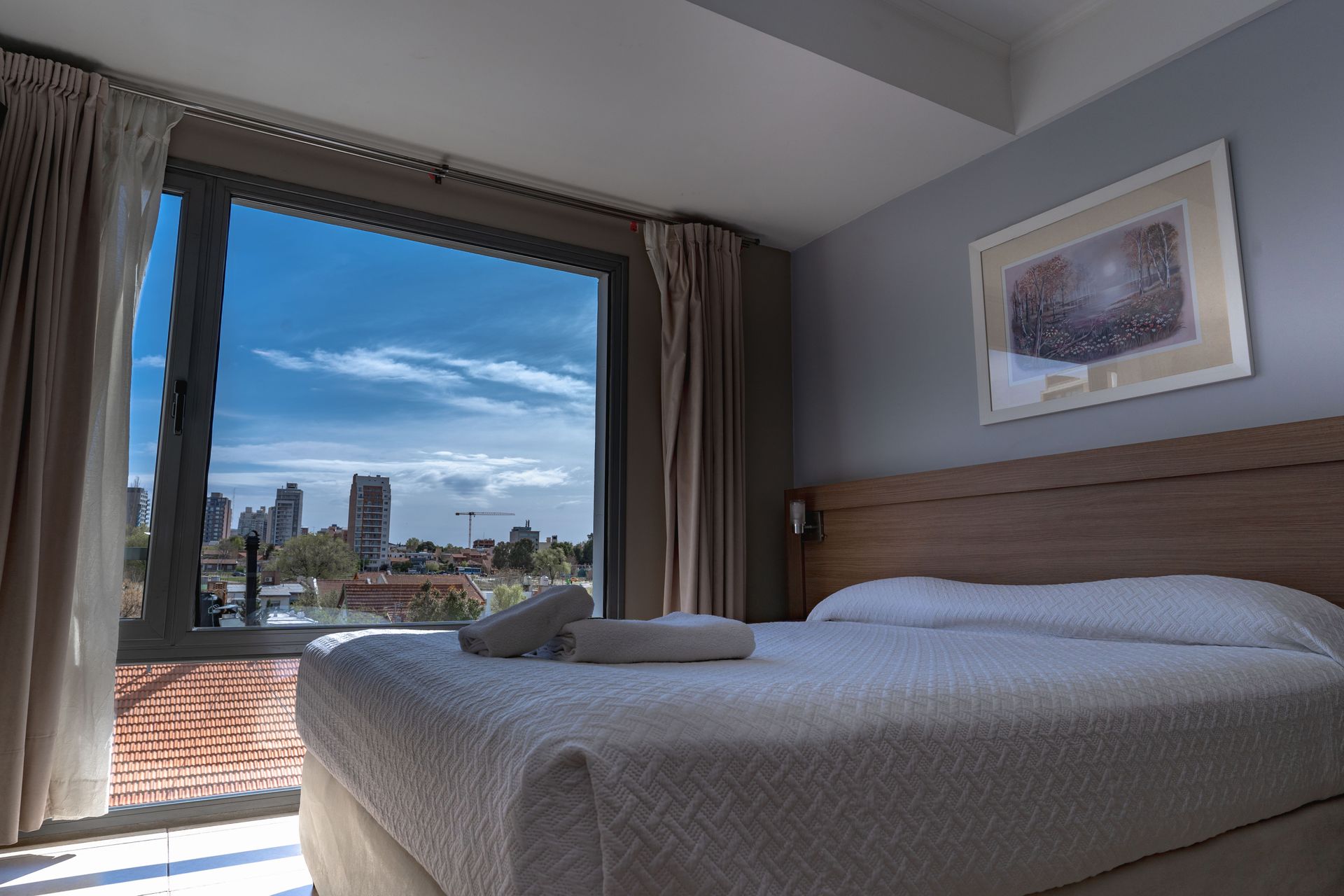 Bedroom with a large window overlooking a city skyline. White bed, gray walls, and a framed picture.