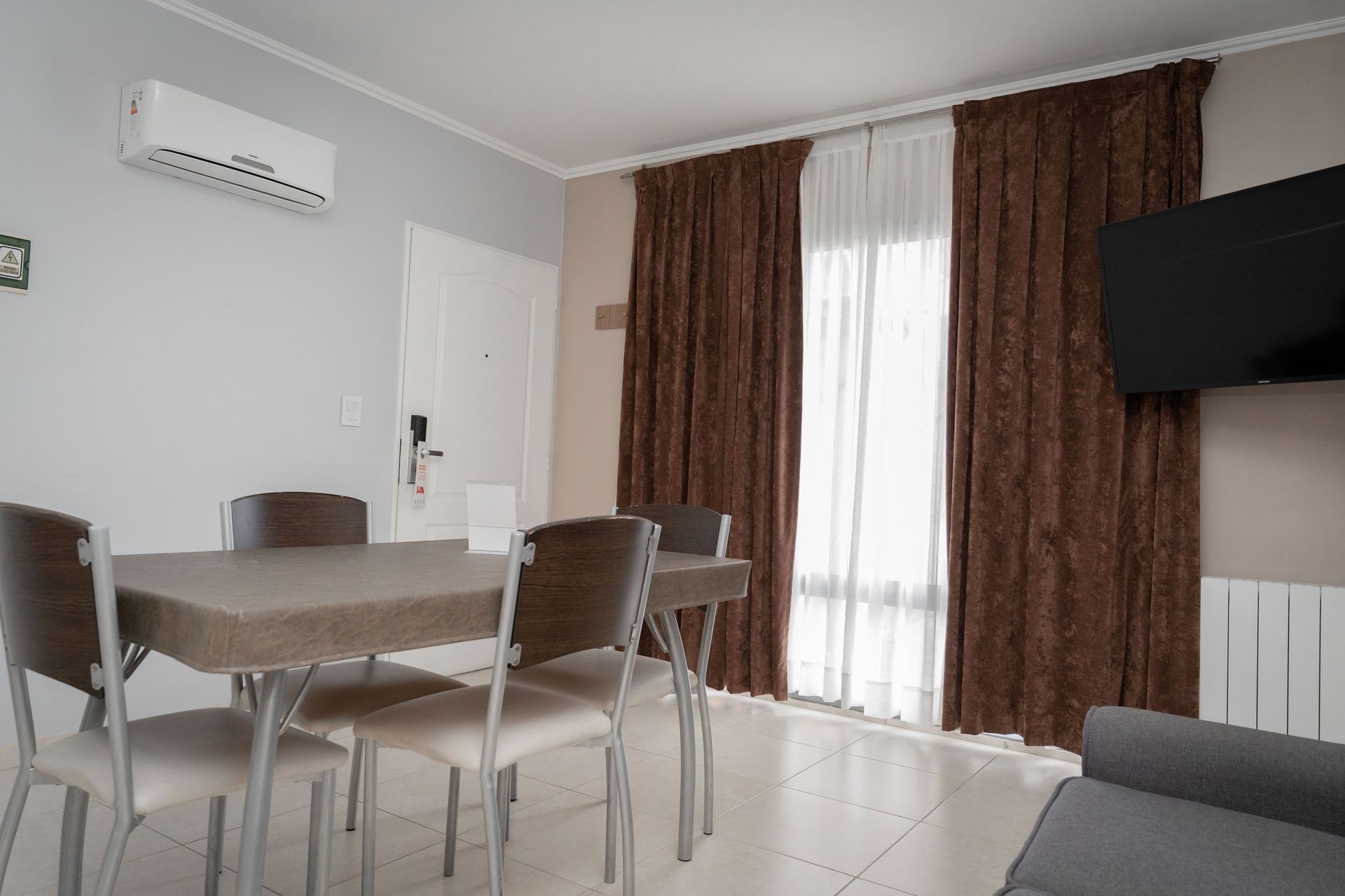 Dining area with table, chairs, TV, window with brown curtains, and a/c unit.