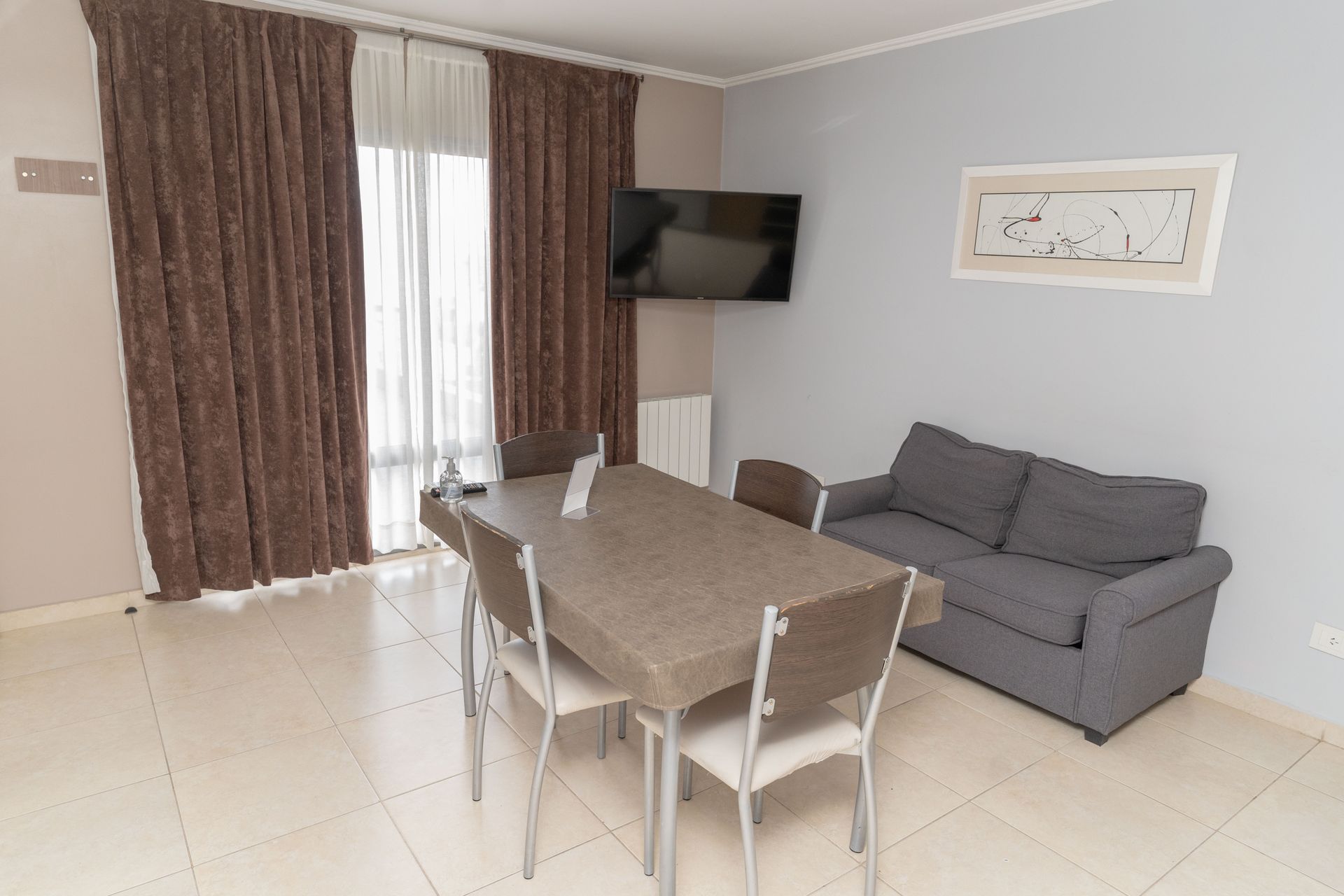 Living room with dining table, sofa, TV, and window with brown curtains. Light-colored walls and floor.