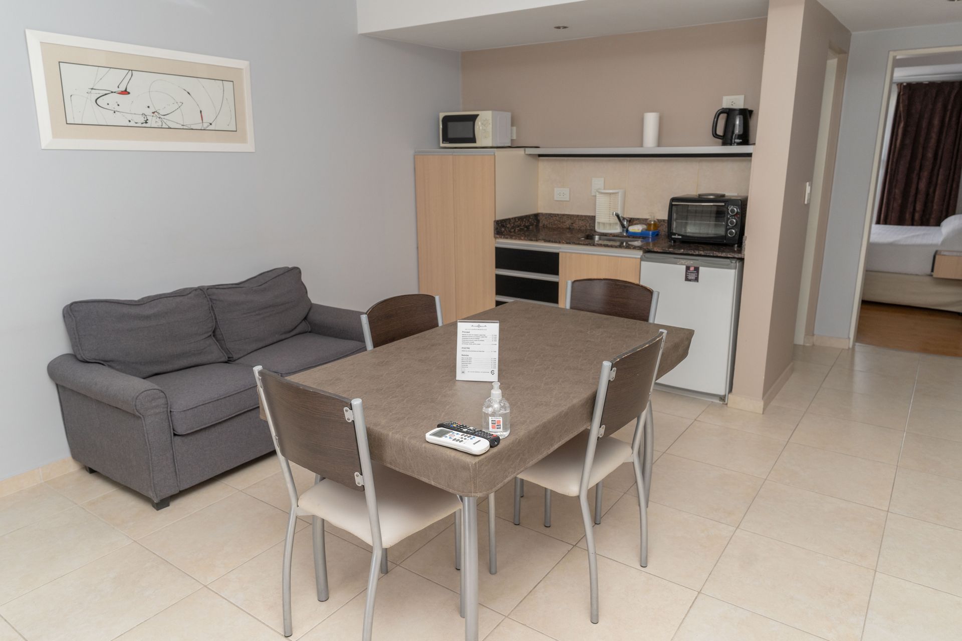 Studio apartment with a gray sofa, dining table, kitchenette, and open doorway to bedroom.