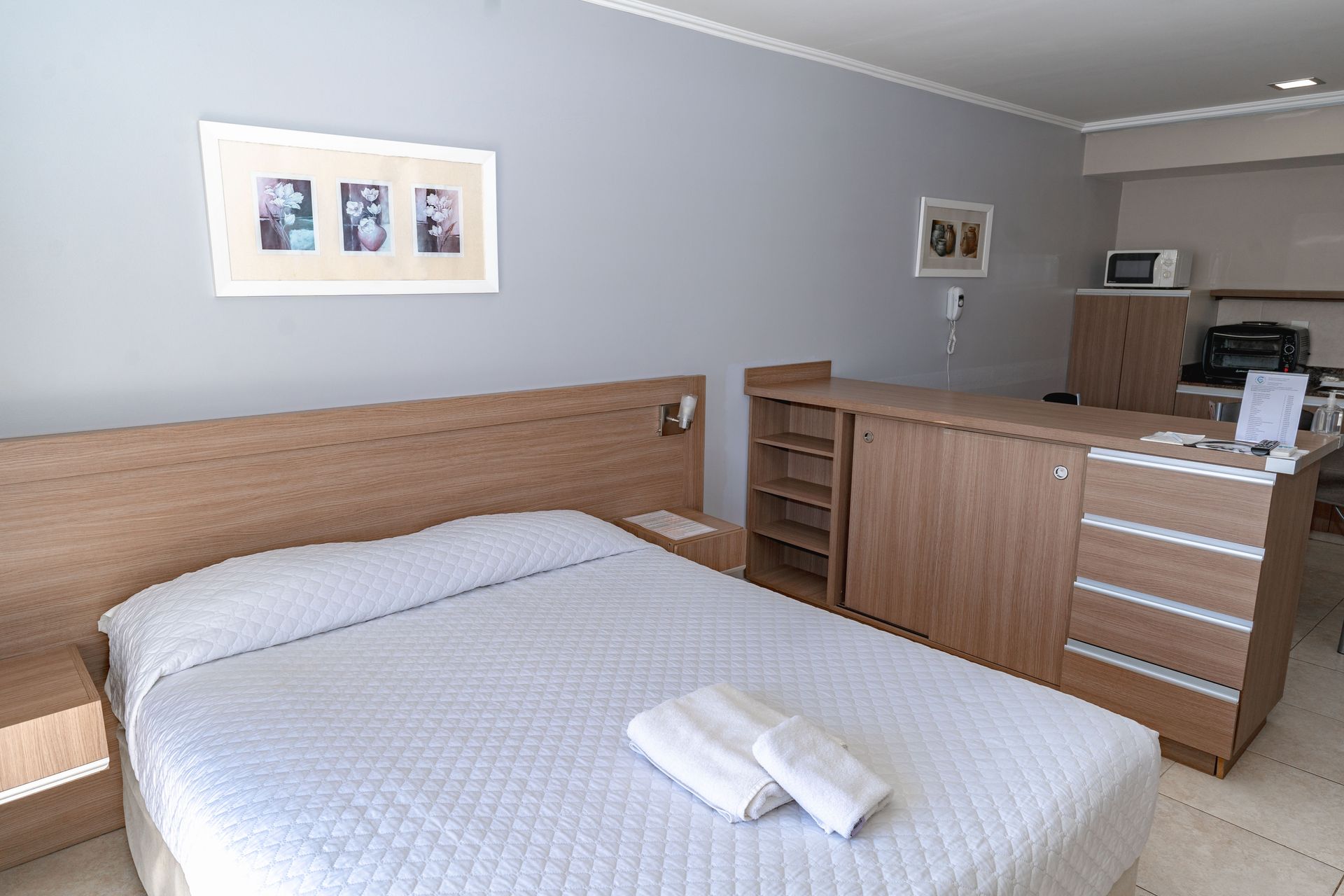 Bedroom with a bed, nightstands, built-in desk, cabinets, microwave, and framed artwork. Light wood and gray color scheme.