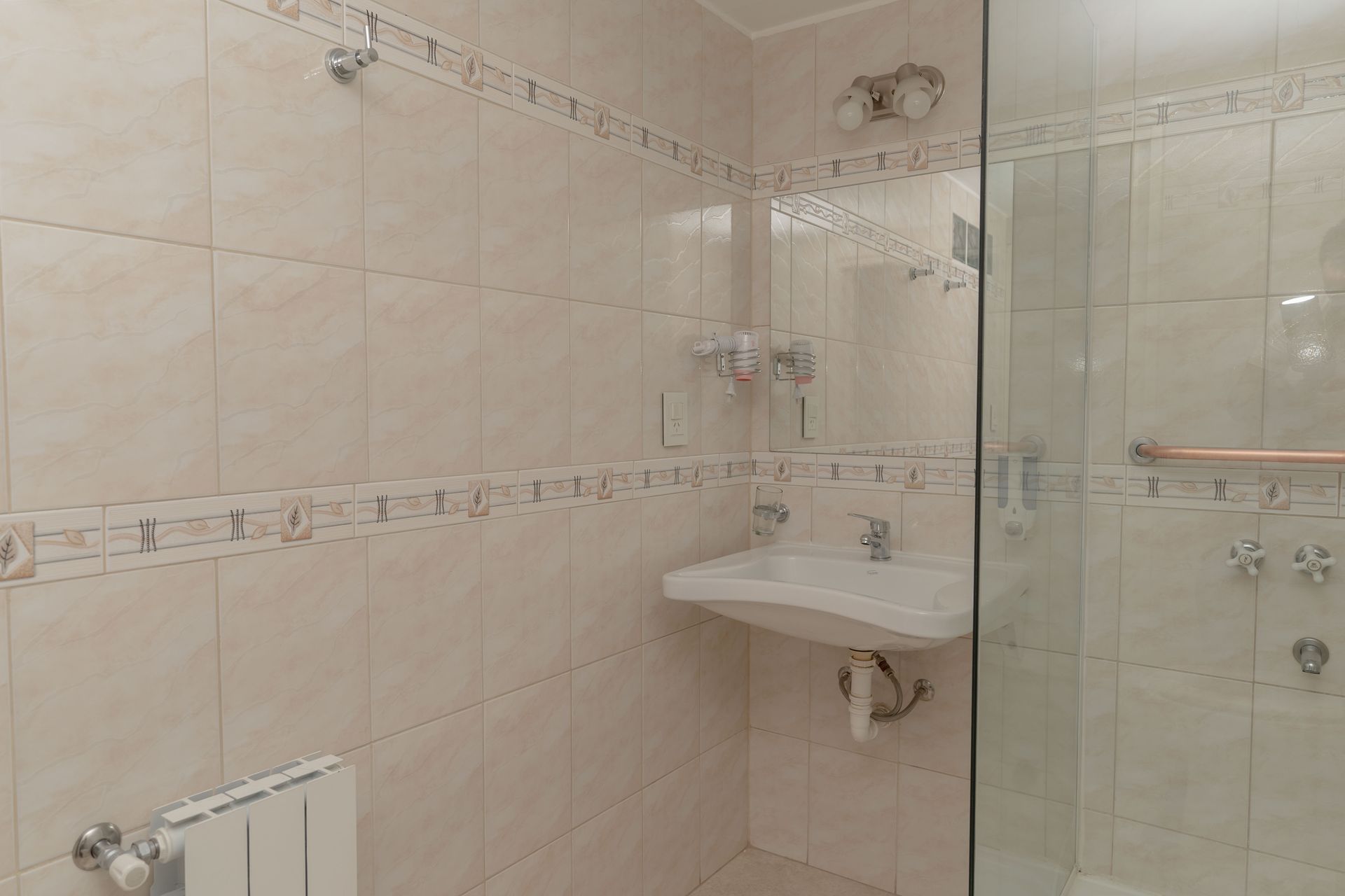 Bathroom with beige tiled walls, a sink, a mirror, and a glass shower door.