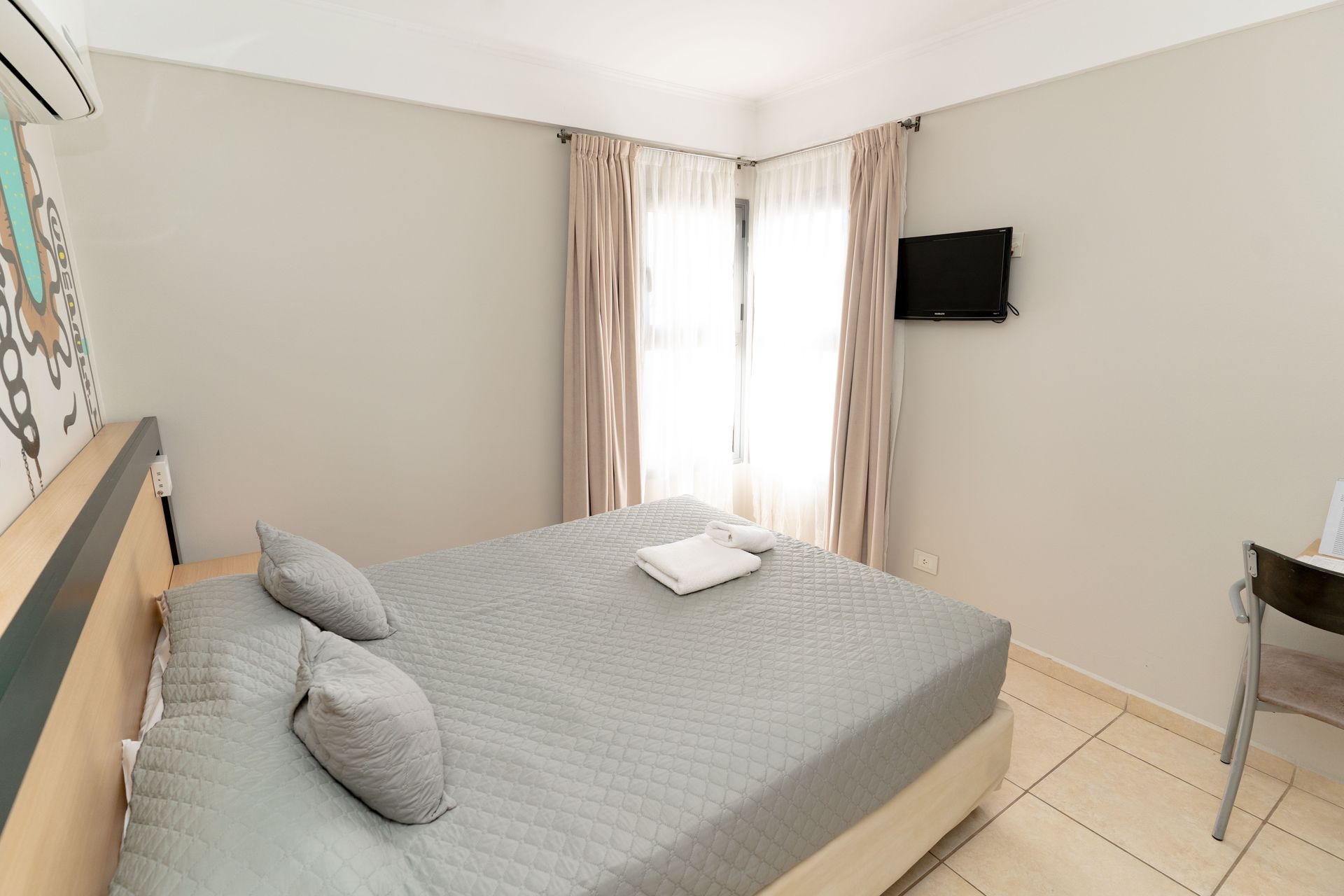 Bedroom with a bed, pillows, a window, a TV, and a desk. The walls are light grey.