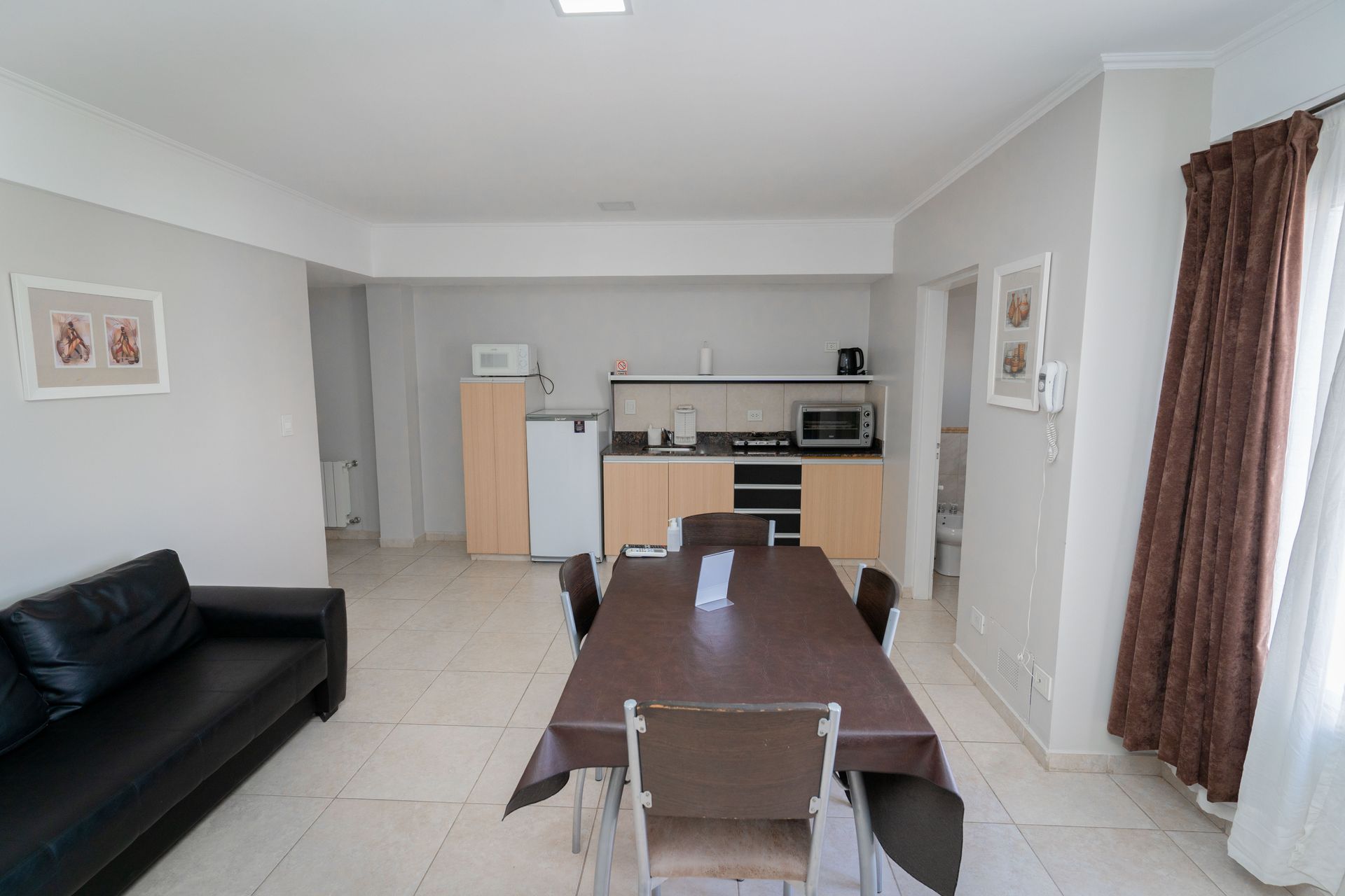 Living area with kitchen, dining table, and black sofa. Beige walls, brown curtains.
