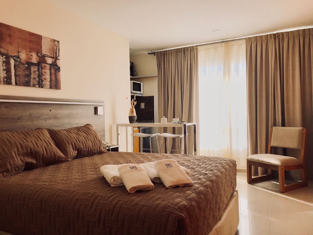 Hotel room with a bed, desk, chair, and curtains. Earth-tone decor.