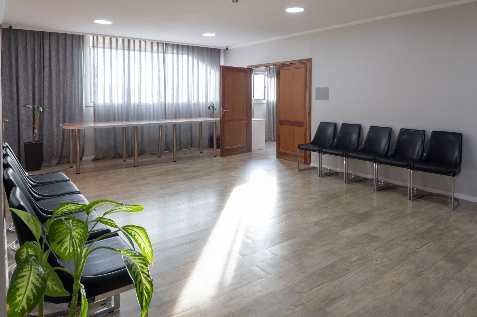 Empty waiting room with black chairs, long table, and a plant. Light streams in from a window.