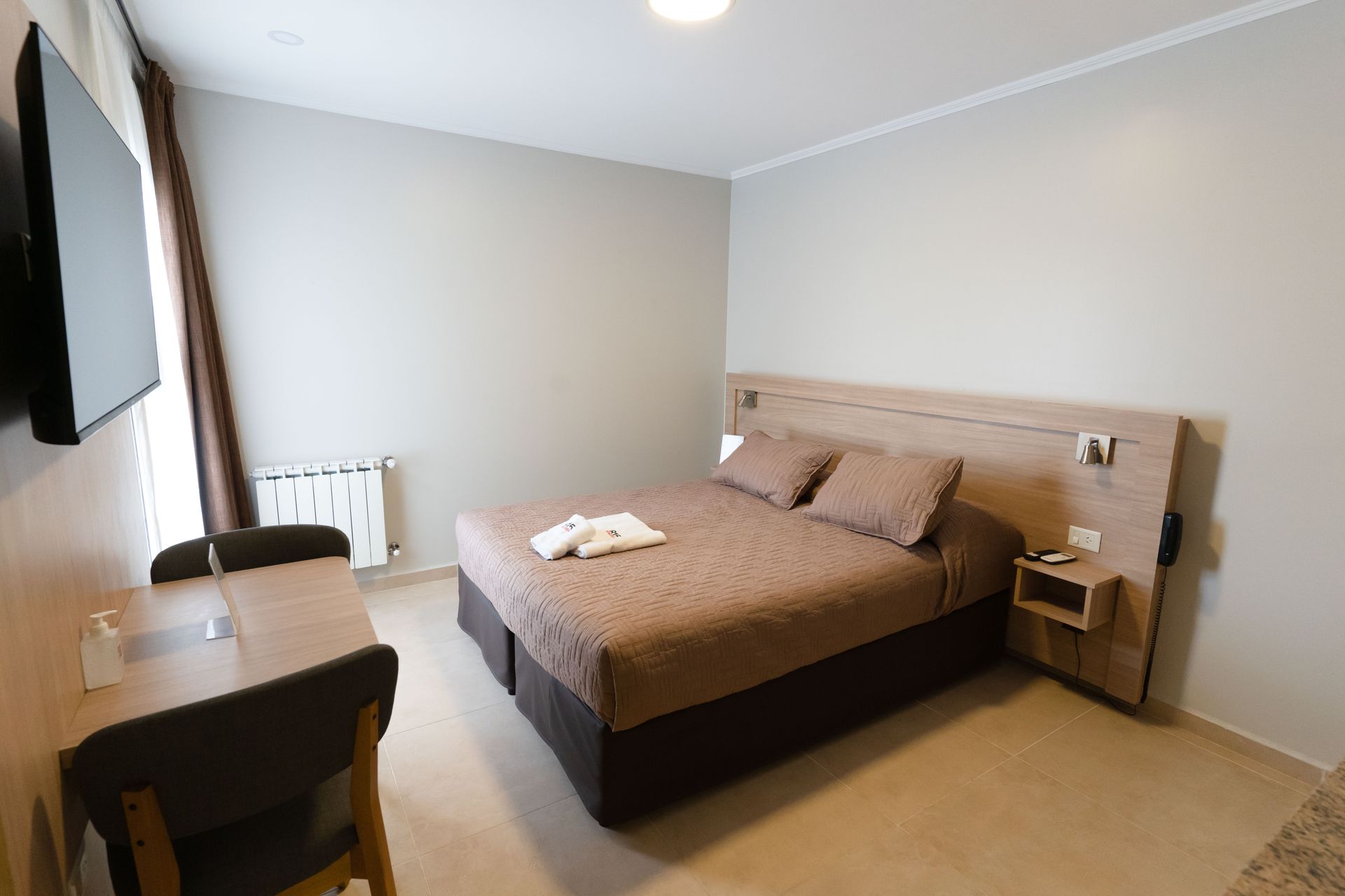 Hotel room with a bed, desk, and TV; light brown and beige tones.