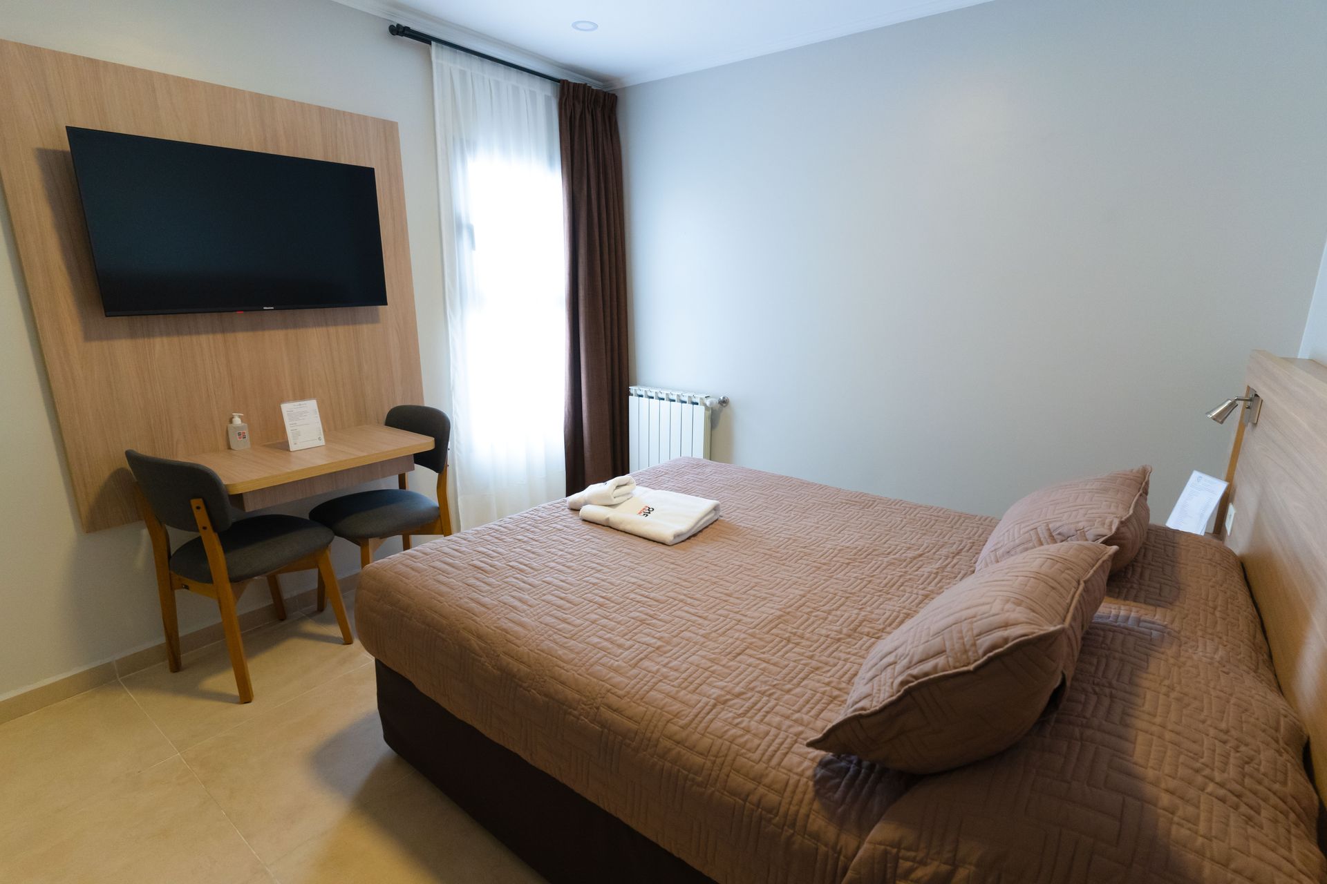 Bedroom with double bed, small table with chairs, TV on wall, window with curtains.