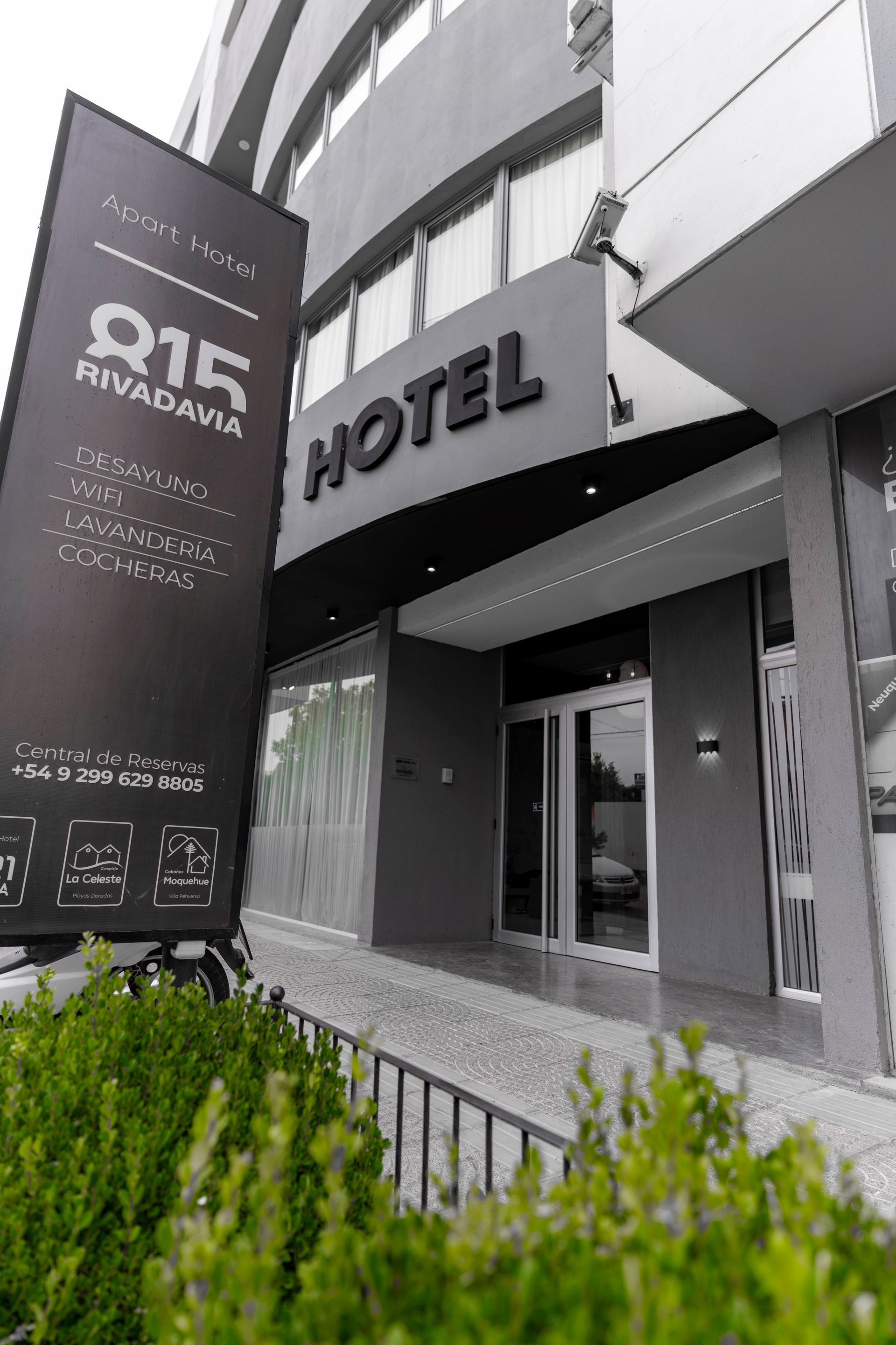 Exterior of a modern white building with parking and a hotel entrance. A sign says 