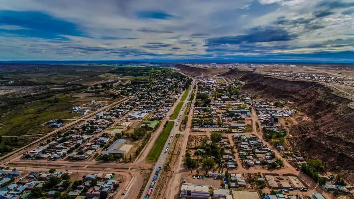 Aerial view of a town with a long road, buildings, and a dramatic canyon on the right, under a cloudy sky.
