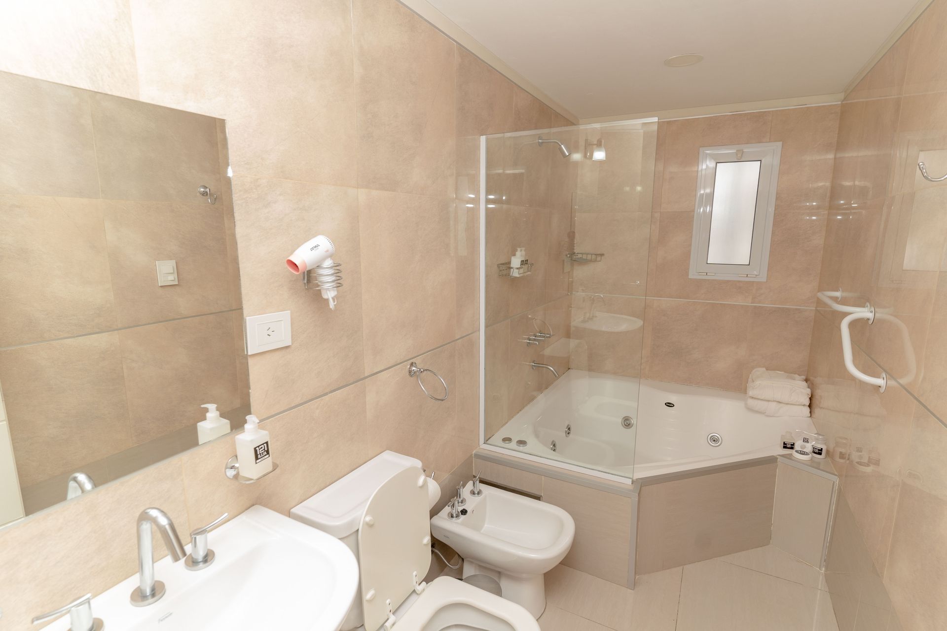Bathroom with a jacuzzi tub, toilet, and sink; beige tiled walls and a small window.