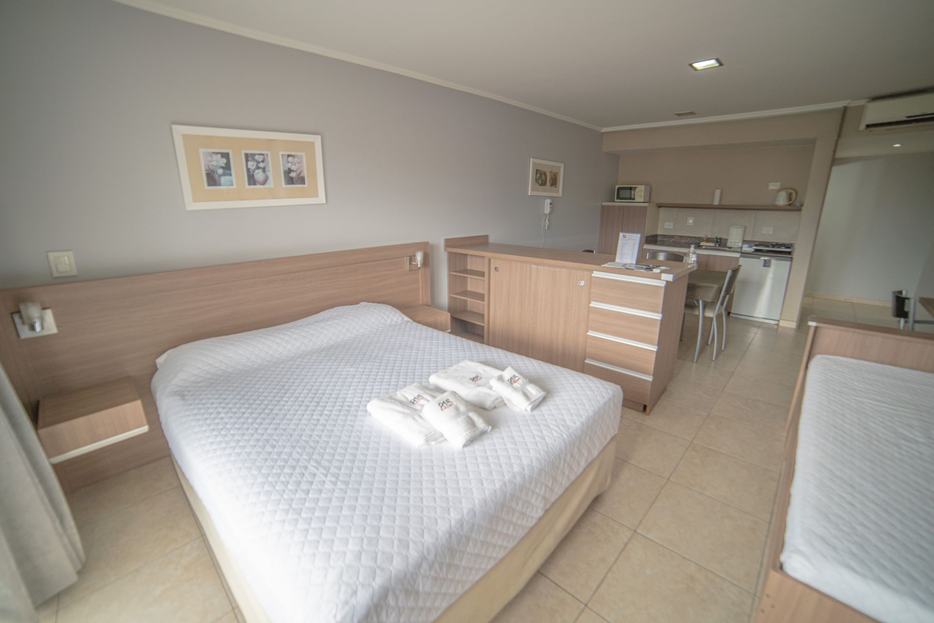 Hotel room with a bed, kitchenette, and a second bed. Light-colored walls and furniture.