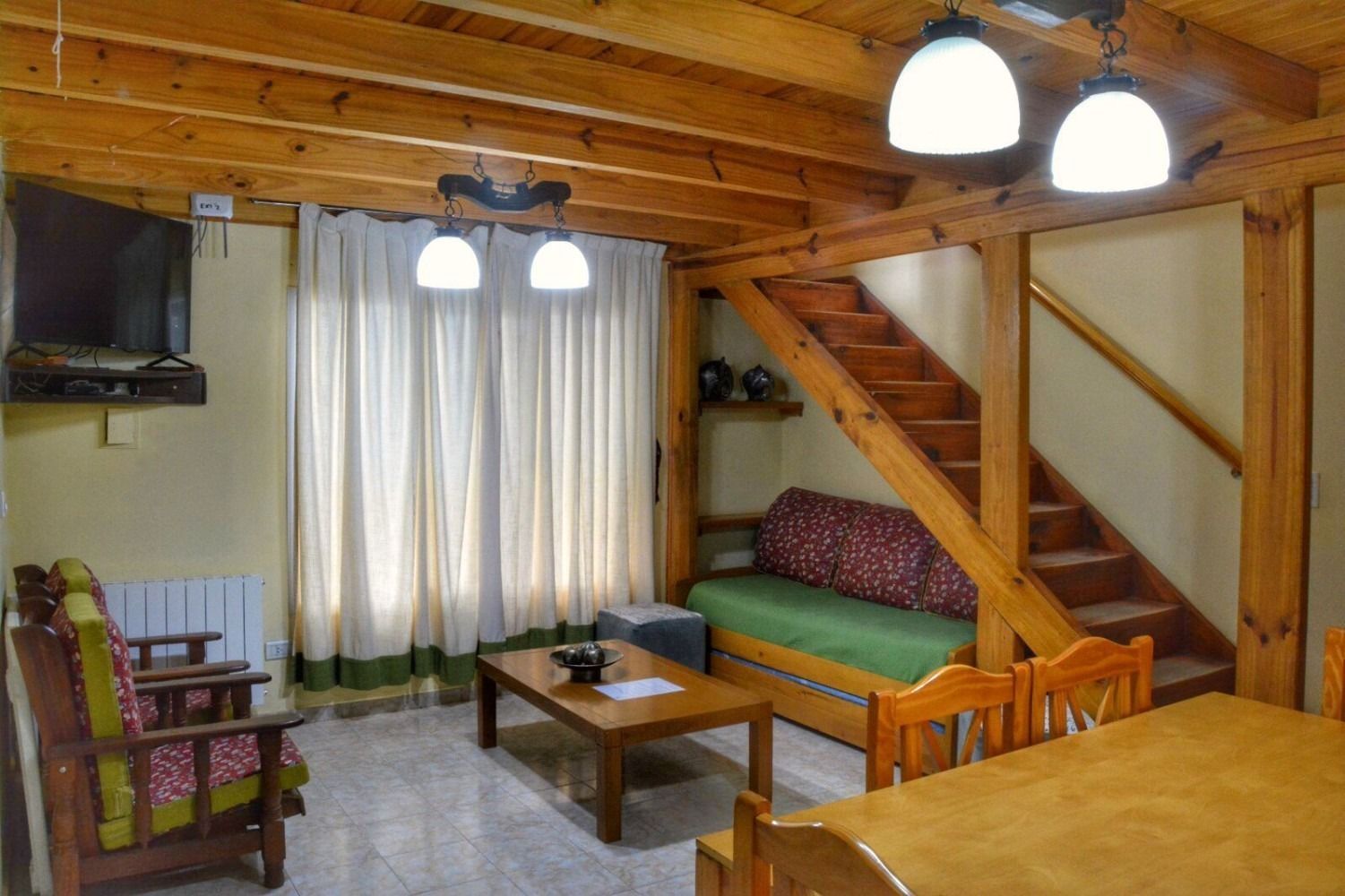 Cozy cabin living room with wooden beams, stairs, and furniture; includes couch, table, and television.