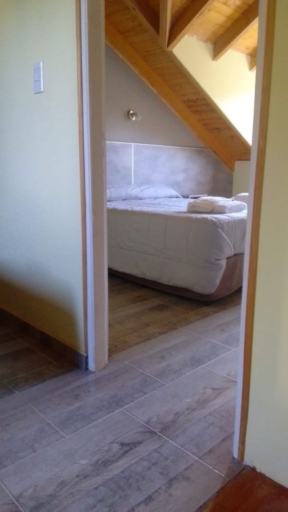 View through a doorway into a bedroom with a bed. The room has a slanted ceiling, wood beams, and tile floor.