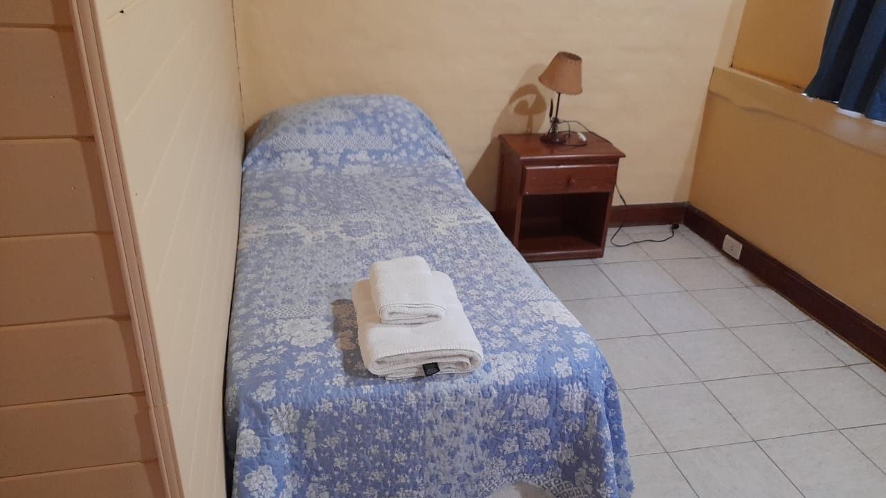 A small, simple bedroom with a single bed covered with a blue floral patterned blanket and a nightstand with a lamp.