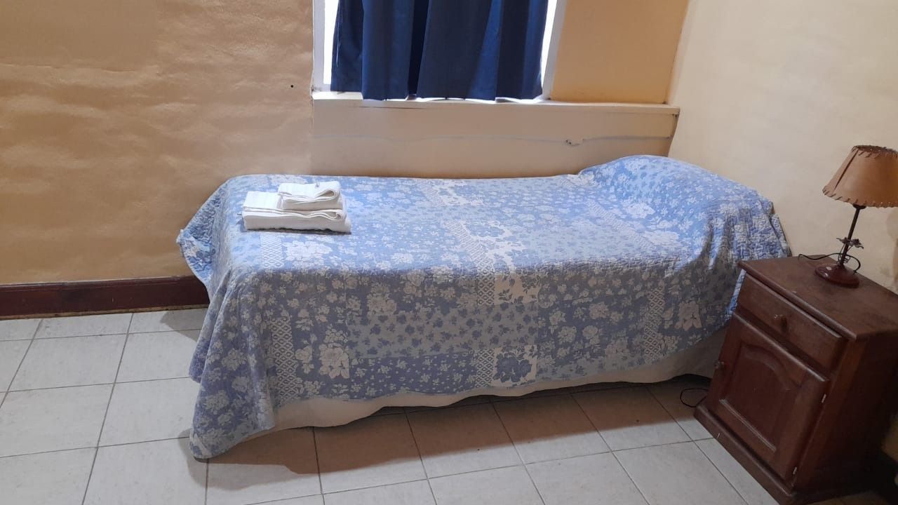 A single bed with a blue and white patterned bedspread, nightstand, lamp, and small window.