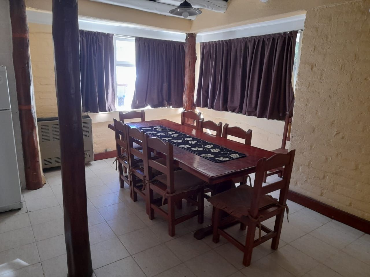 Dining room with long wooden table and chairs, brown curtains, and exposed beams.