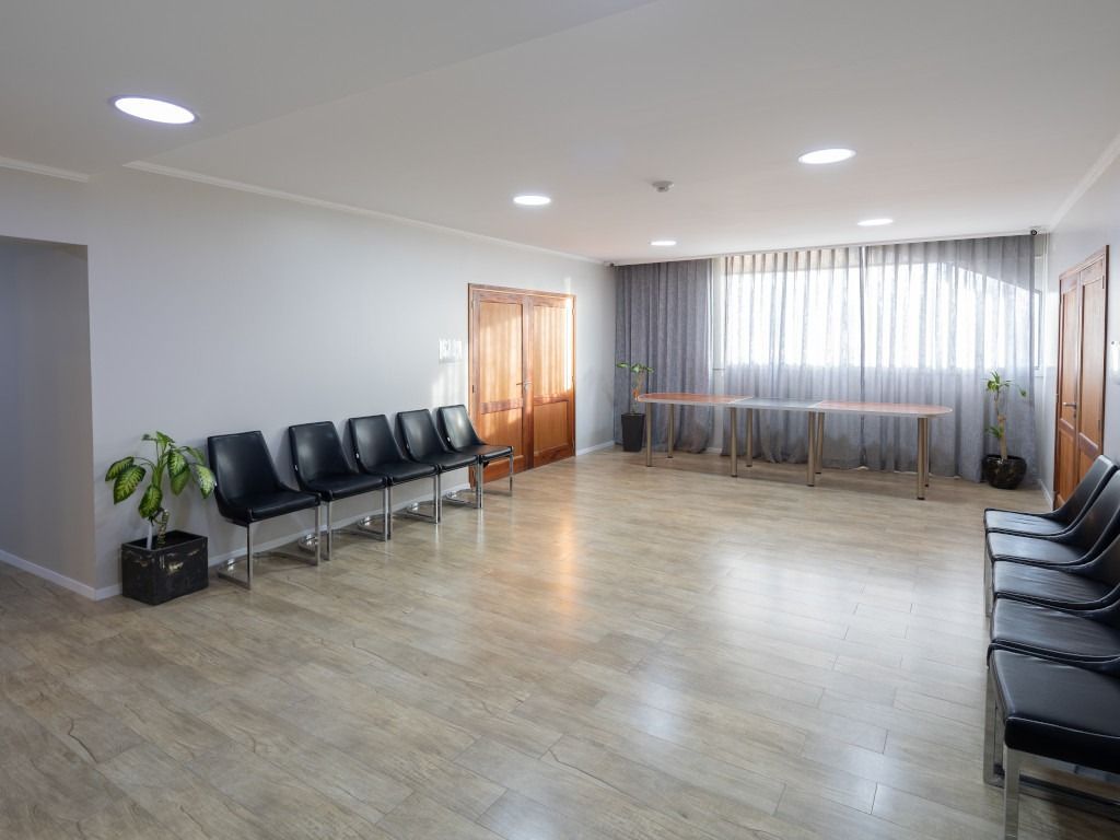 Empty waiting room with black chairs, a long table, and a window with sheer curtains.