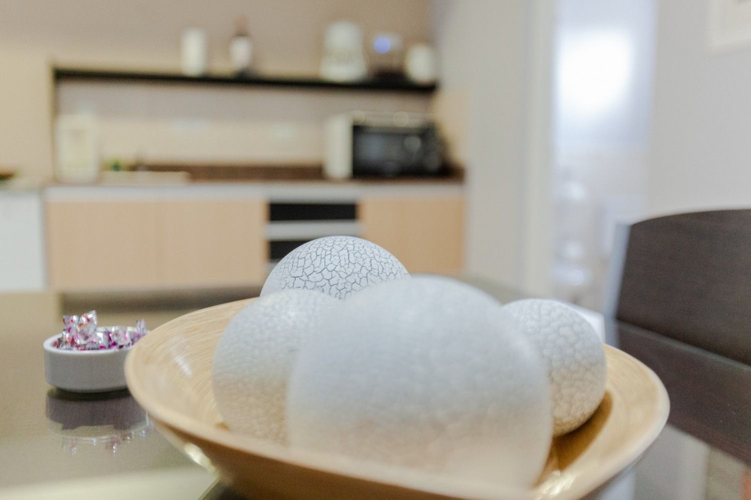 A wooden bowl with decorative white spheres on a table, with a blurred kitchen in the background.
