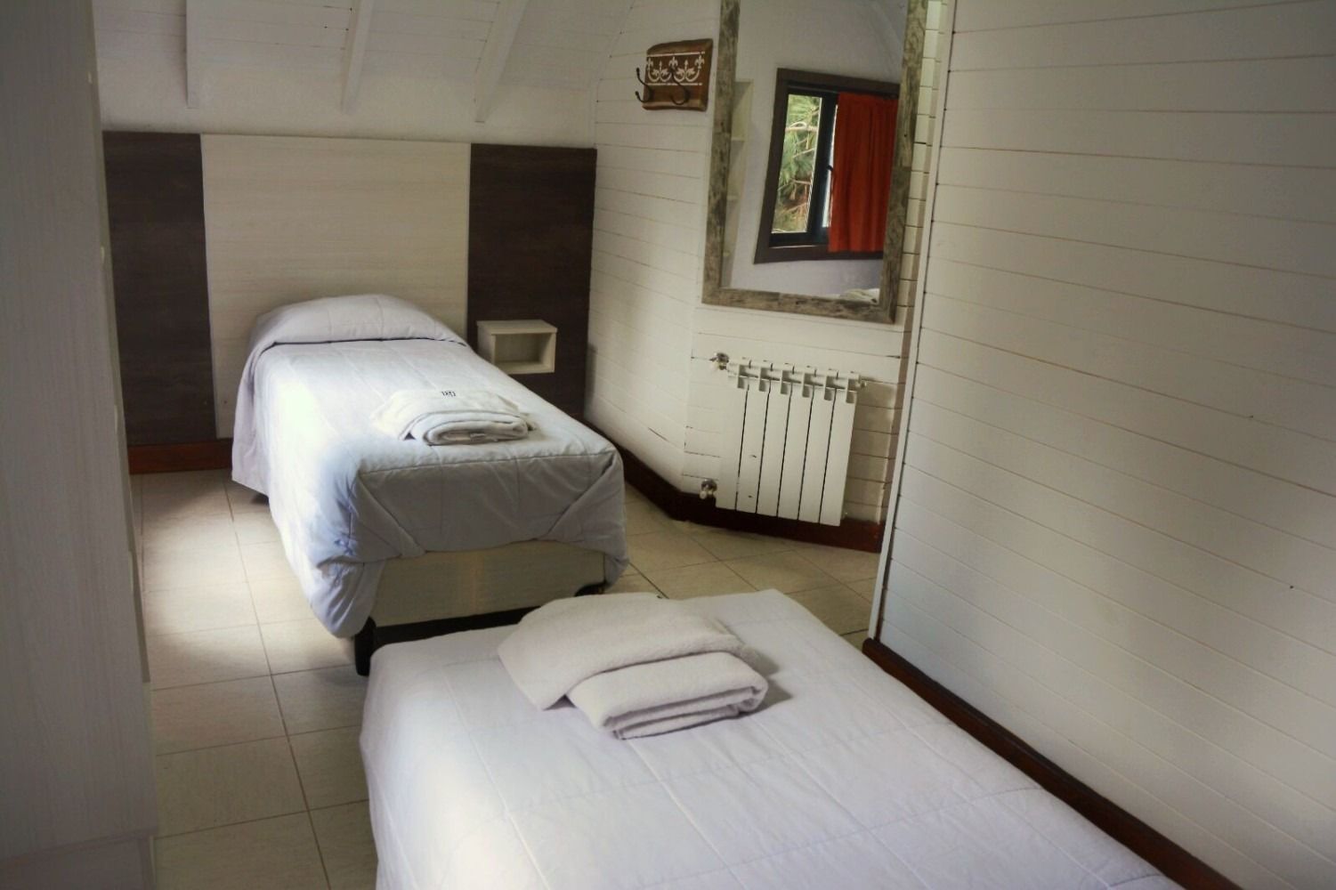 Two twin beds in a white-walled room with a window, mirror, and radiator.