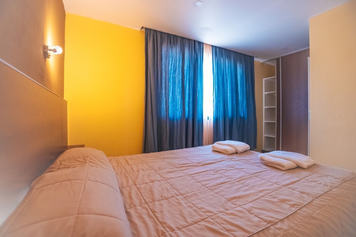 Bedroom with bed, yellow and gray walls, blue curtains, and white towels.