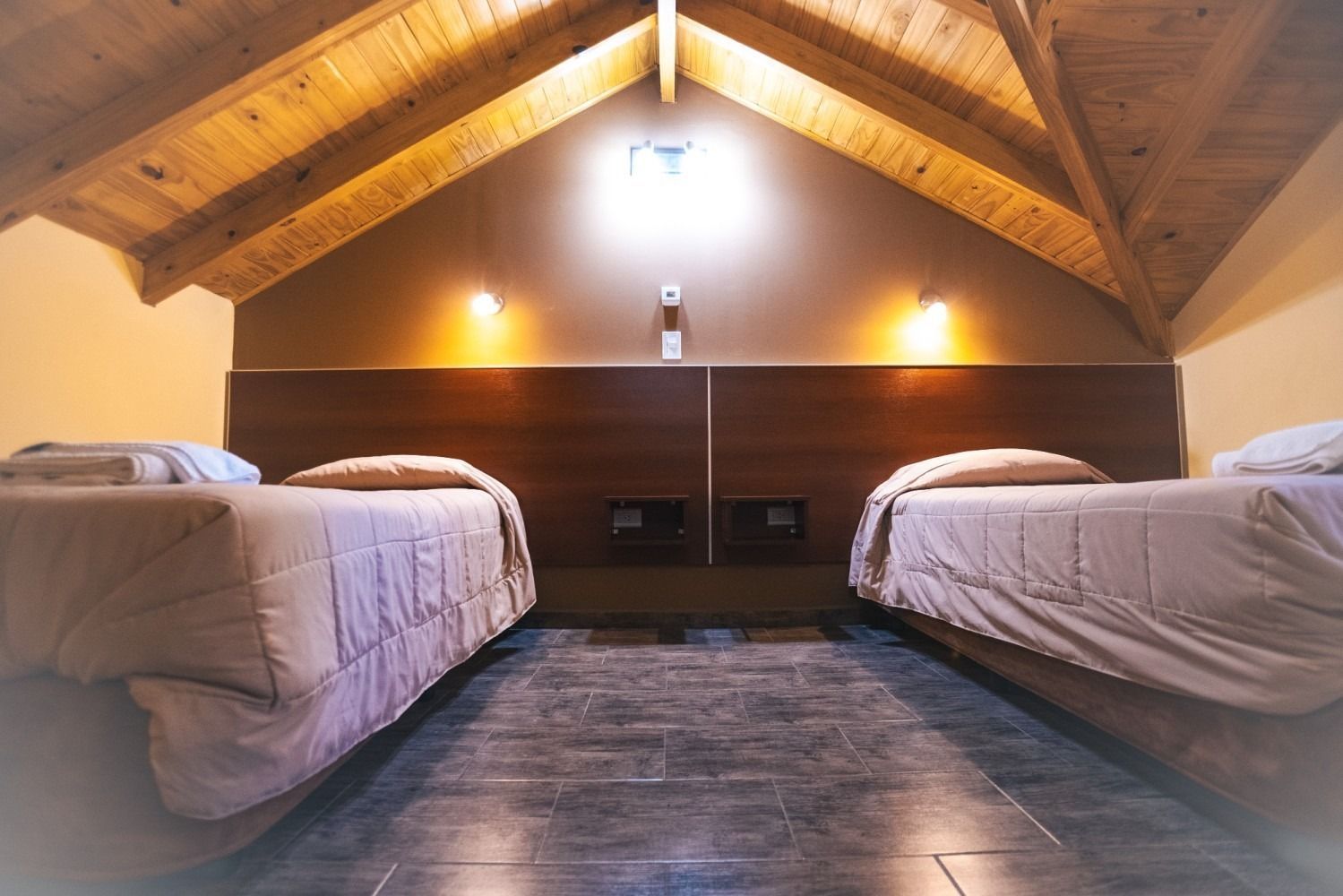 Two beds in a room with a sloped wooden ceiling. Dark brown headboard, neutral bedding.