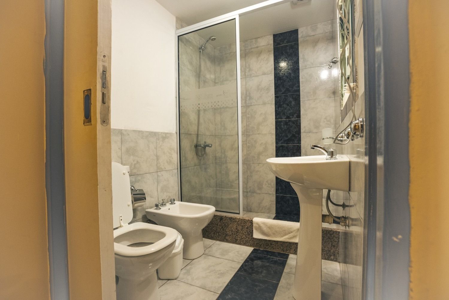 Bathroom with a toilet, bidet, sink, and shower stall. Gray tiled walls and floor, and glass shower door.
