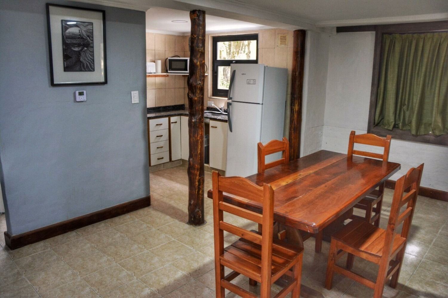 Dining area with wooden table, chairs, and a kitchen visible in the background. Light-colored walls and flooring.