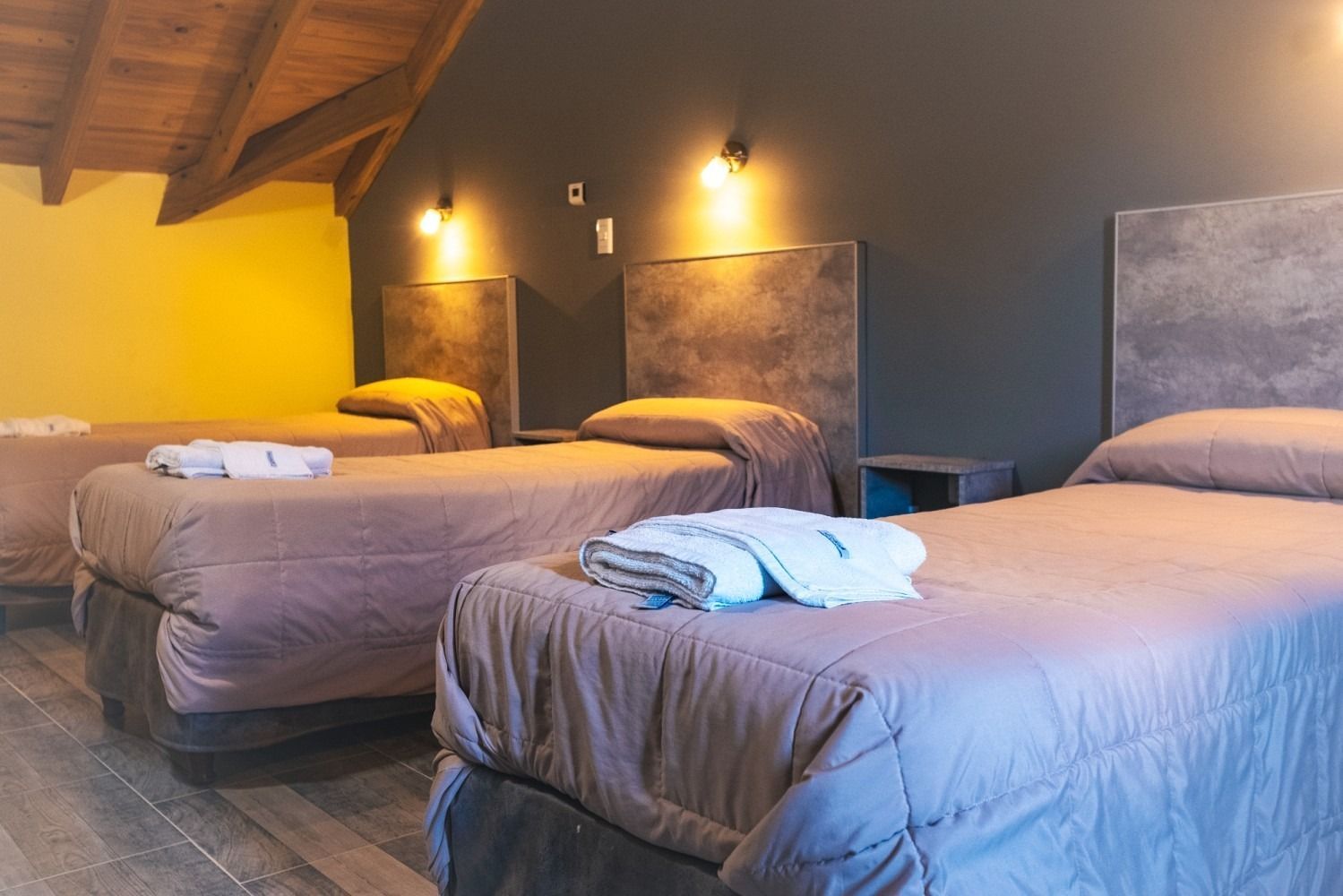 Three beds in a room, brown bedding, dark gray wall, wood ceiling, soft lighting.