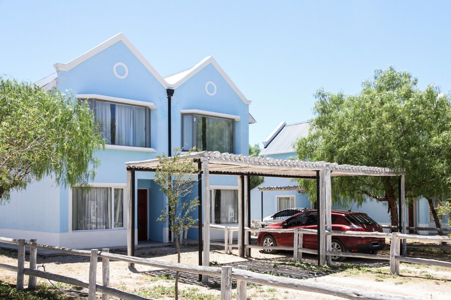 Light blue house with white trim, a car under a pergola, trees, and a wooden fence.