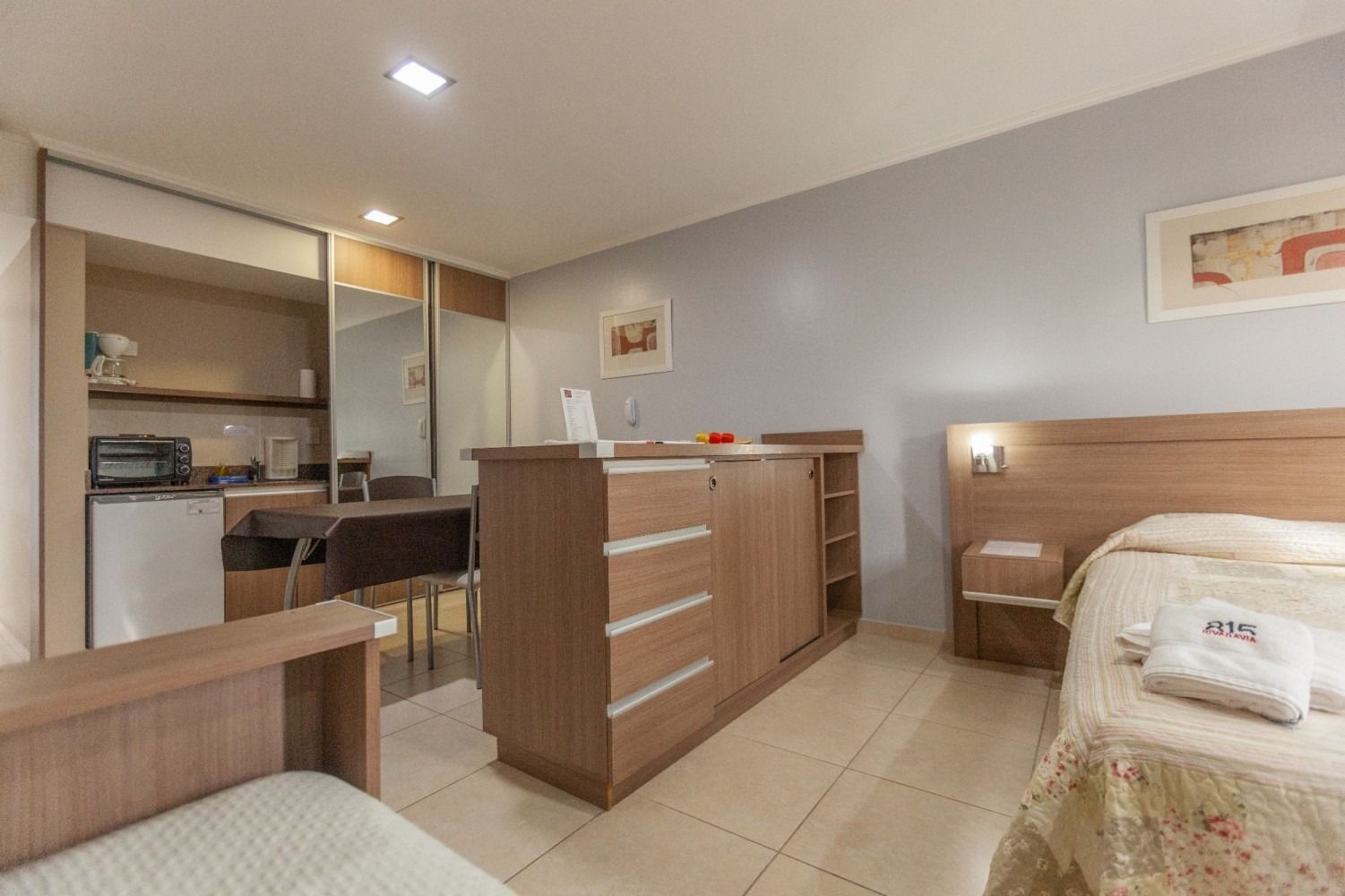 Studio apartment interior with kitchen, bed, and sitting area. Light wood and neutral colors.