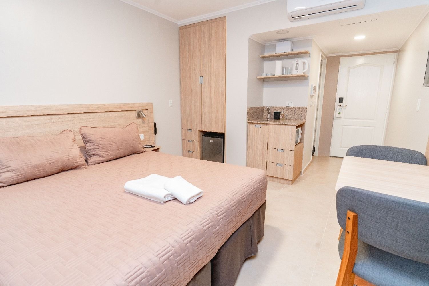 Bedroom with bed, closet, kitchenette, and dining table. Neutral colors and minimal decor.
