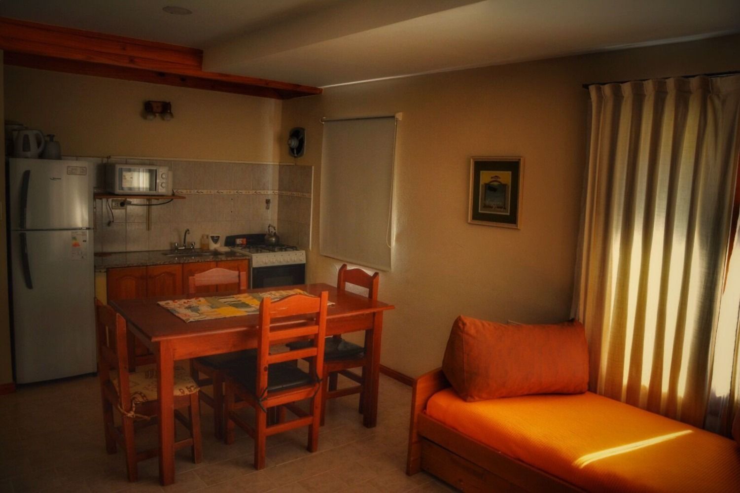 Cozy living space with kitchen and dining area; features refrigerator, microwave, wooden table and chairs, and orange daybed.