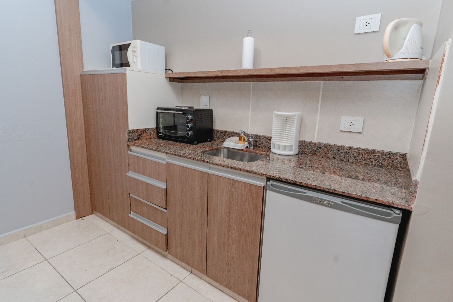 Small kitchenette with cabinets, sink, microwave, refrigerator, and shelf with water boiler.