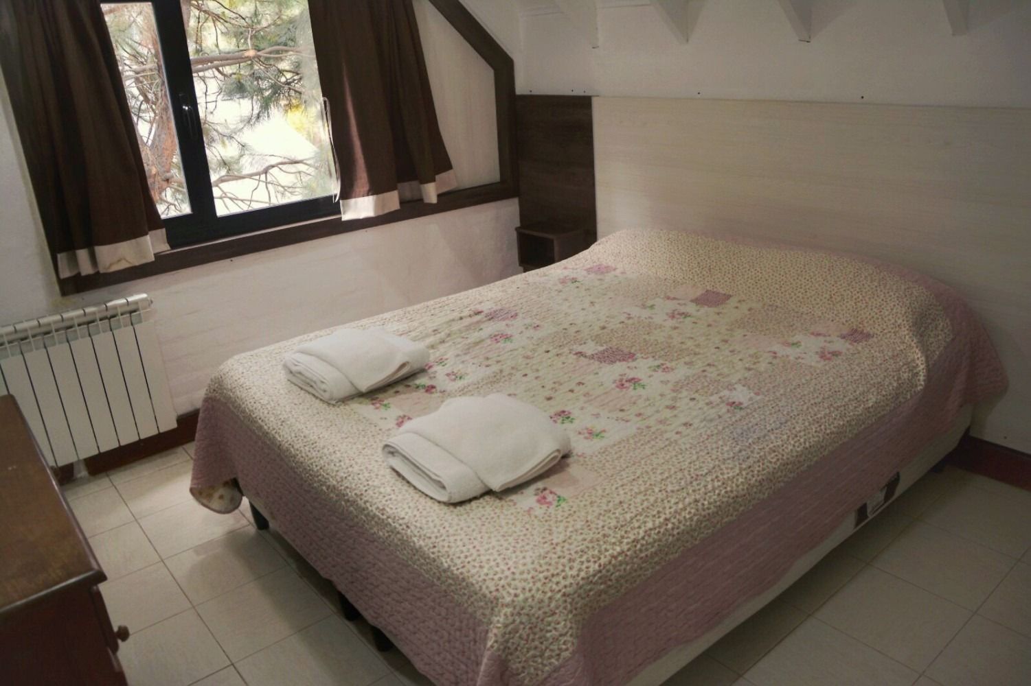 Bedroom with double bed, floral quilt, two folded towels, window with curtains, radiator.