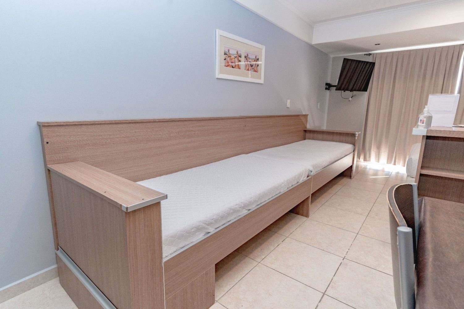 A light-colored wooden daybed against a light blue wall in a room, with a small TV and beige curtains.