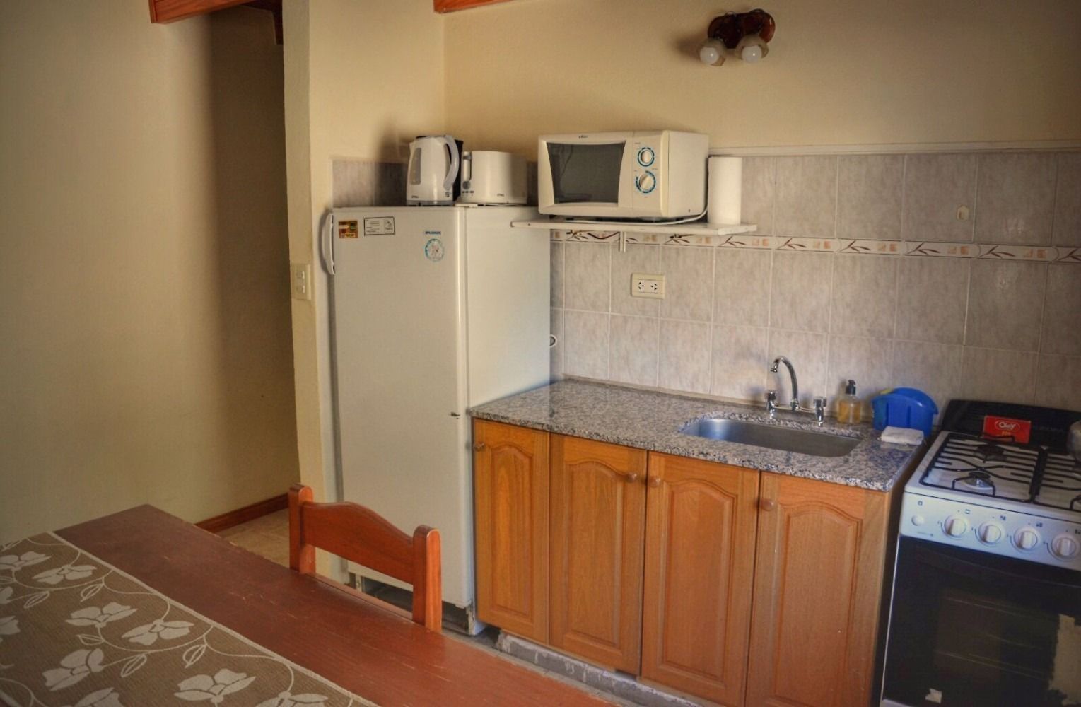 Small kitchen with refrigerator, microwave, sink, and stove. Light brown cabinets and tan walls.