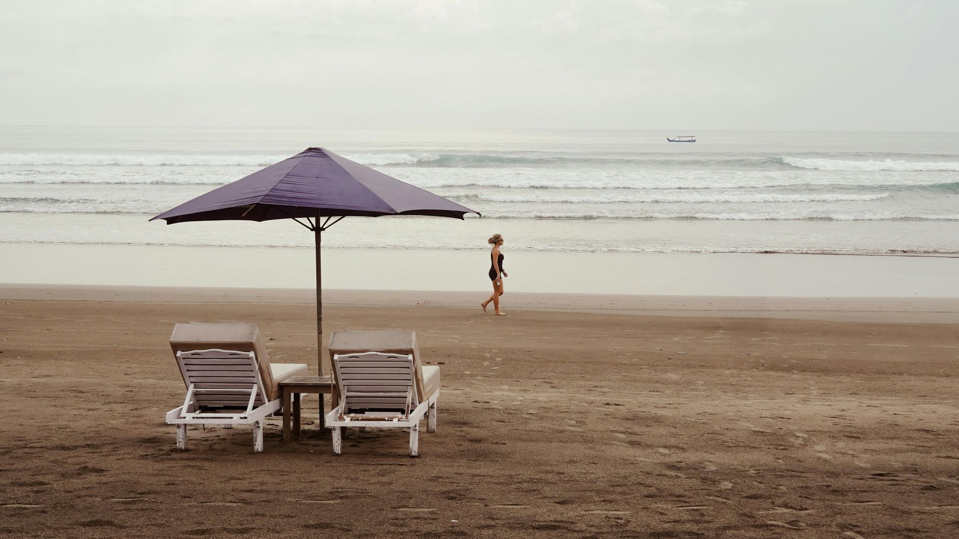 Beach scene: two empty lounge chairs under a purple umbrella, person walking on the wet sand towards the ocean.