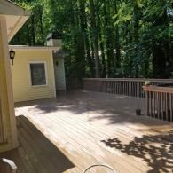 A large, wooden backyard deck attached to a light yellow house, surrounded by a lush green forest.
